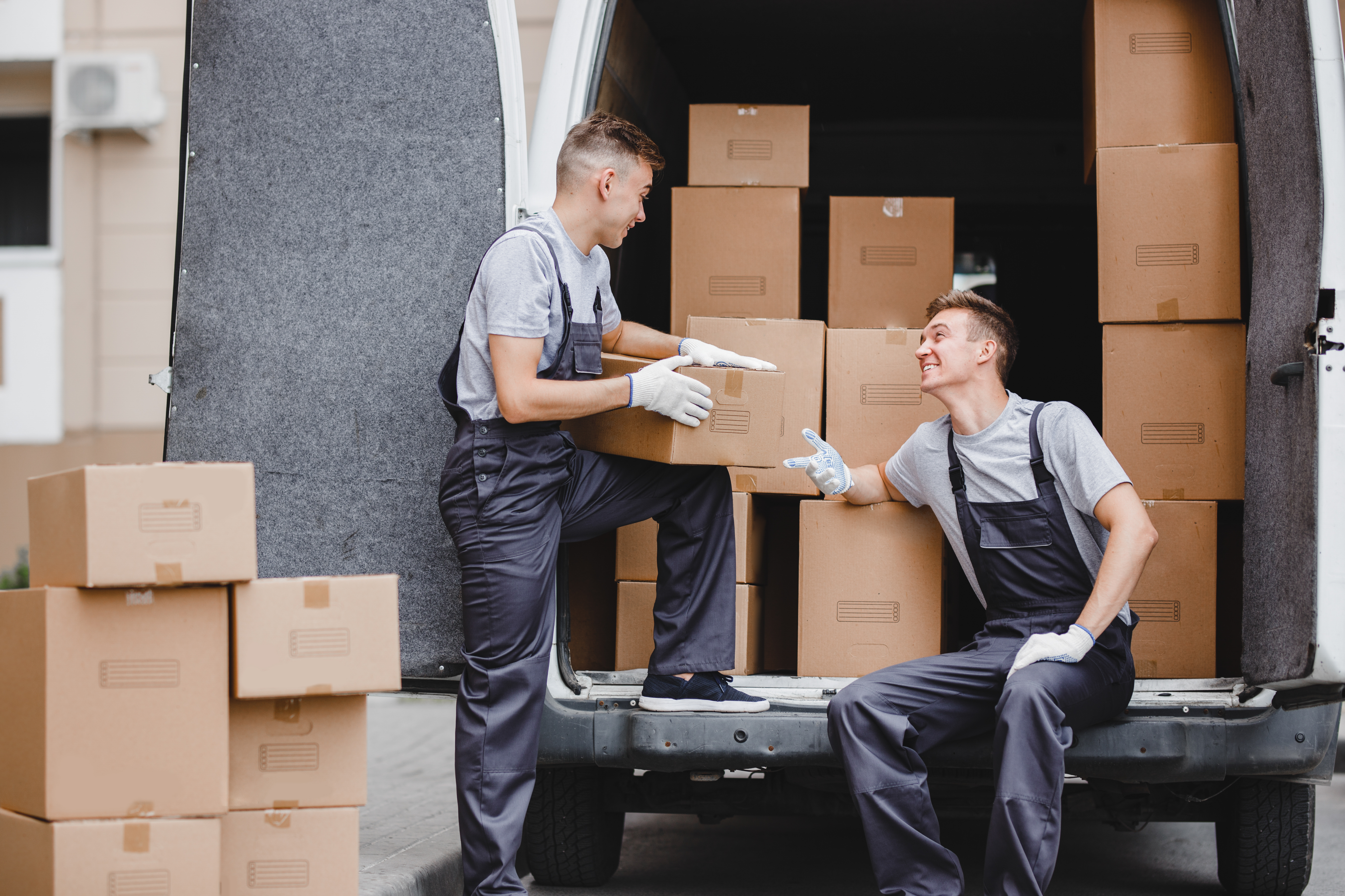 Two young handsome movers wearing uniforms are laughing while unloading the van full of boxes. House move, mover service